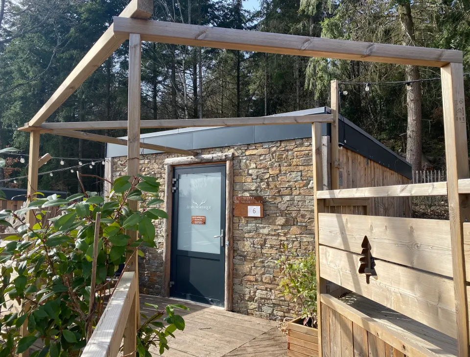 Kleines Steinhaus mit Glastür und der Nummer 6, umgeben von einer Holzkonstruktion und Grünflächen, in einem Wald gelegen